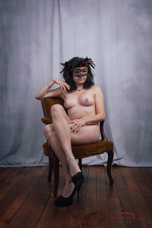 Une jeune femme masquée pose nue en studio assise sur un fauteuil style Louis XV.