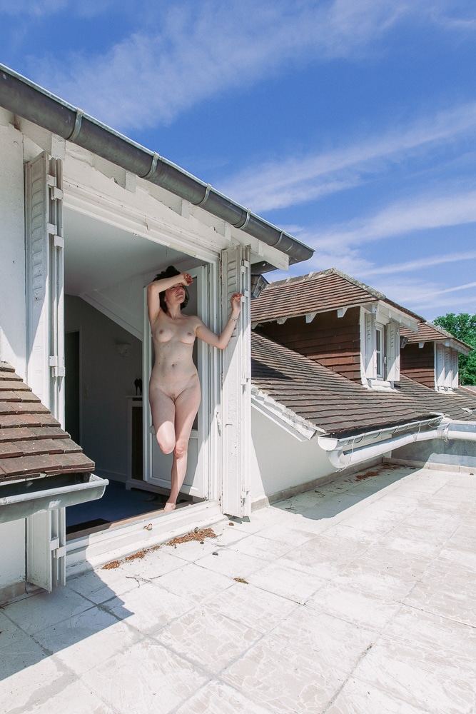 Une femme nue prend la pose dans l'embrasure d'une porte-fenêtre.