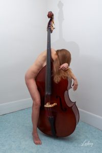 Une femme pose nue avec sa contrebasse dans un angle de pièce.