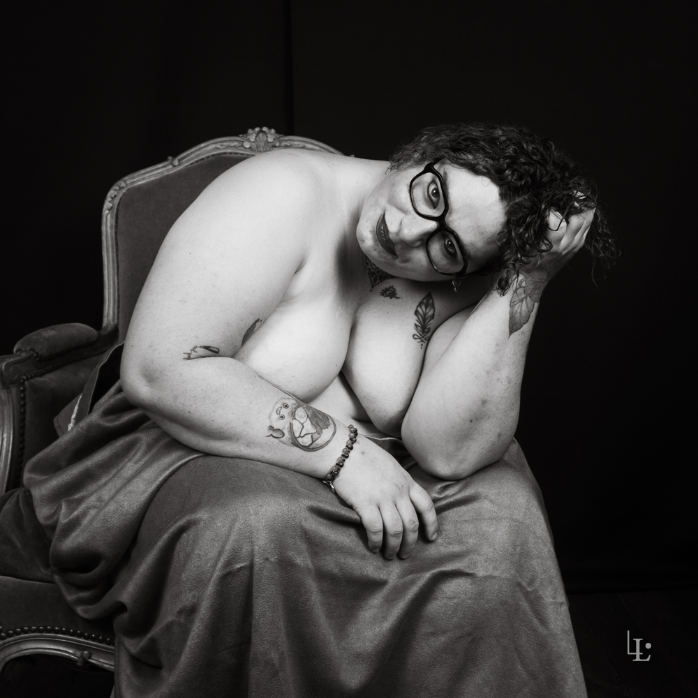 Nu drapé assis sur un fauteuil Voltaire à joues pleines. Photographie studio en noir et blanc.