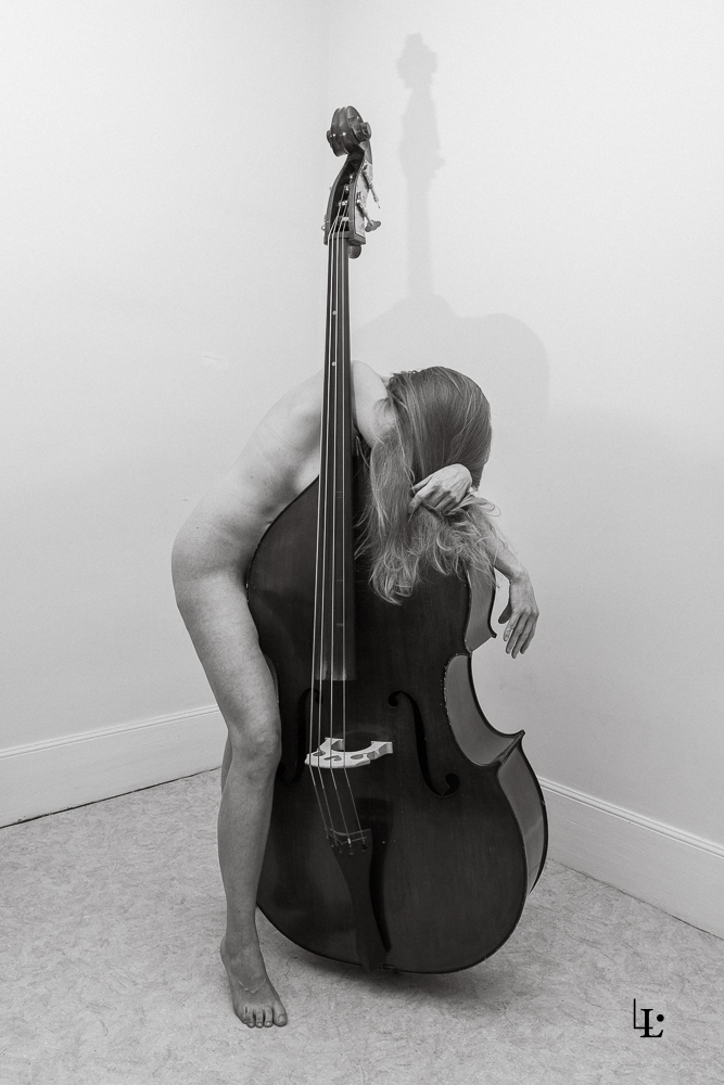 Photographie artistique en noir et blanc montrant une femme nue posant contre une grande contrebasse, le corps épousant l'instrument dans une pose d'abandon.