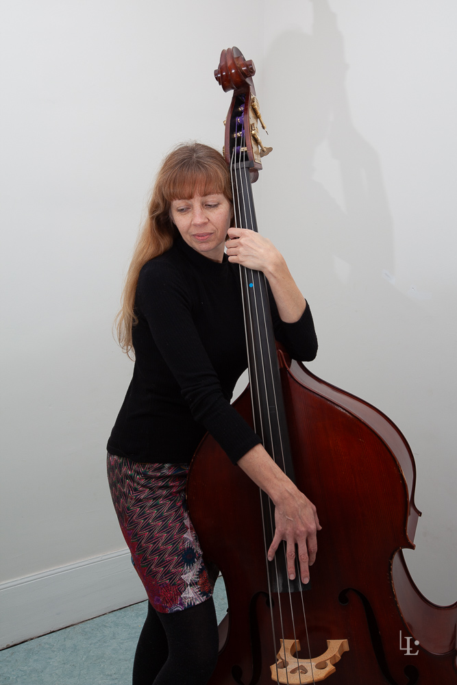 Portrait studio d'une femme posant avec une grande contrebasse, les mains sur l'instrument et le regard baissé, sur un fond blanc.
