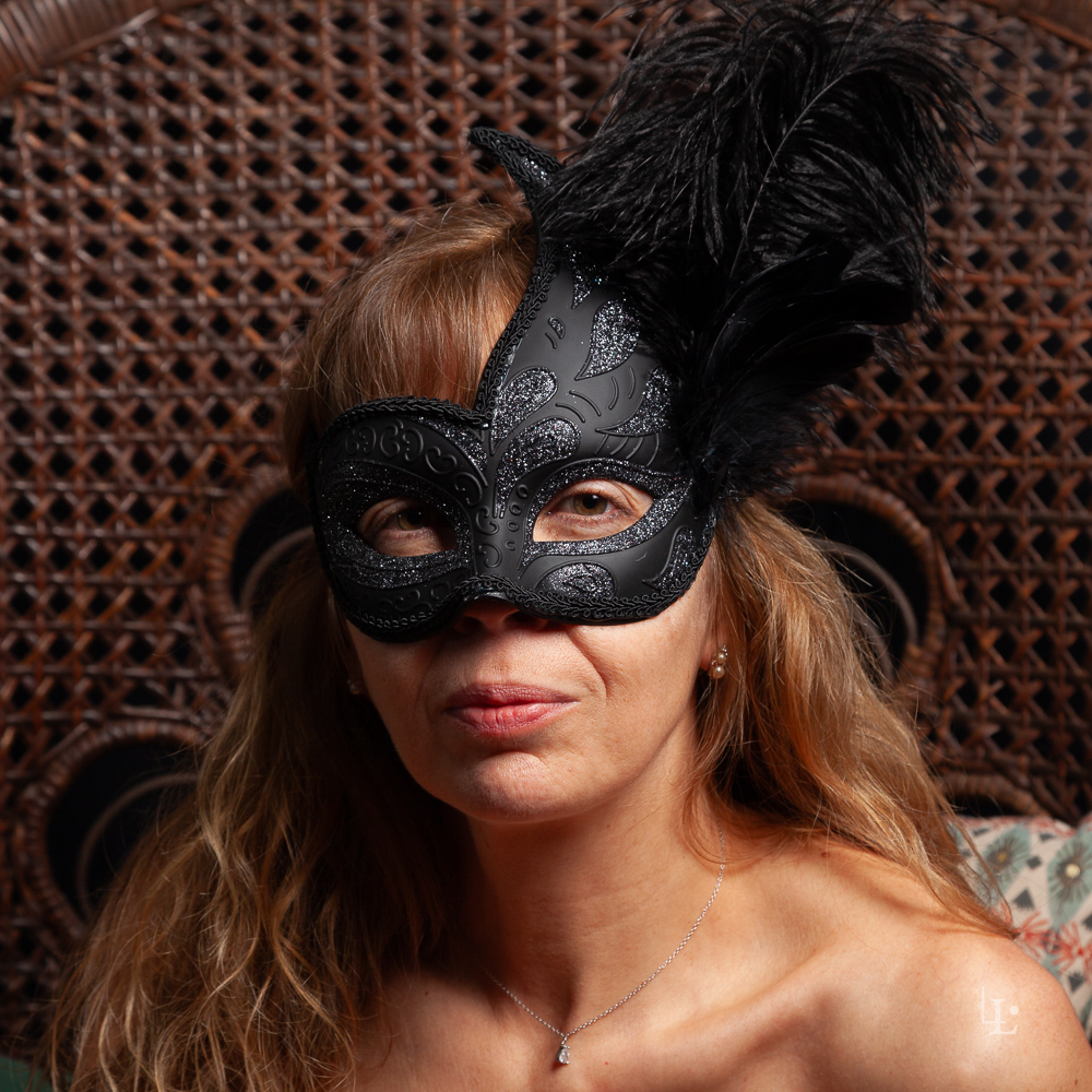 Portrait de femme masquée avec un loup vénitien noir à plumes, assise sur une chaise en rotin.