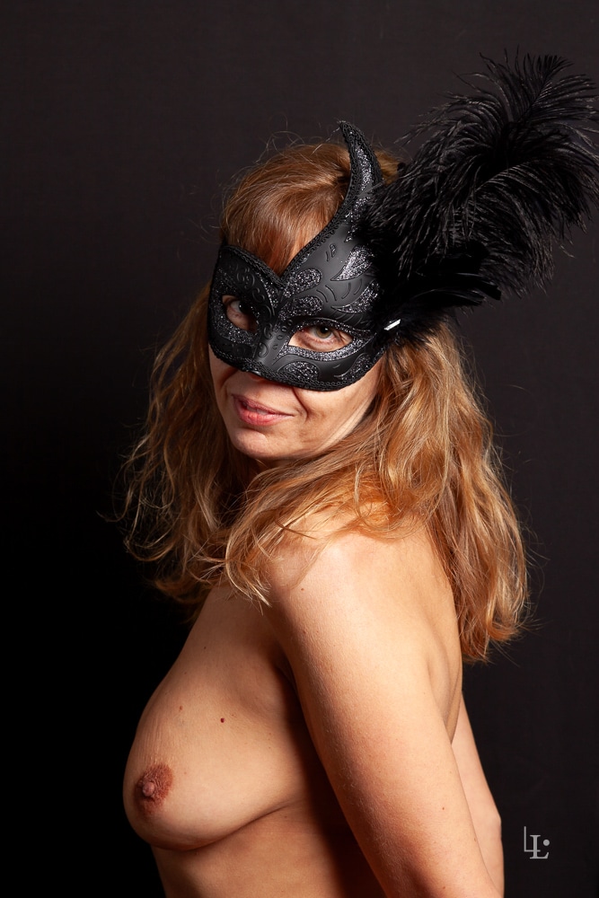Une femme pose en studio de profil le buste nu sous un loup vénitien noir.