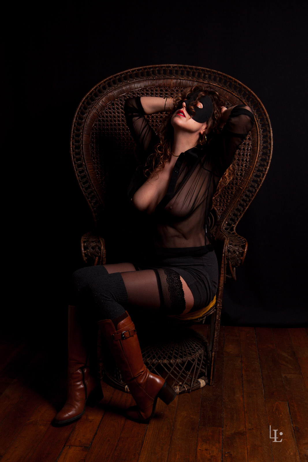 Une jeune femme prend la pose sur un fauteuil Emmanuelle. Elle porte des bottes, un masque ainsi qu'un chemisier transparent.