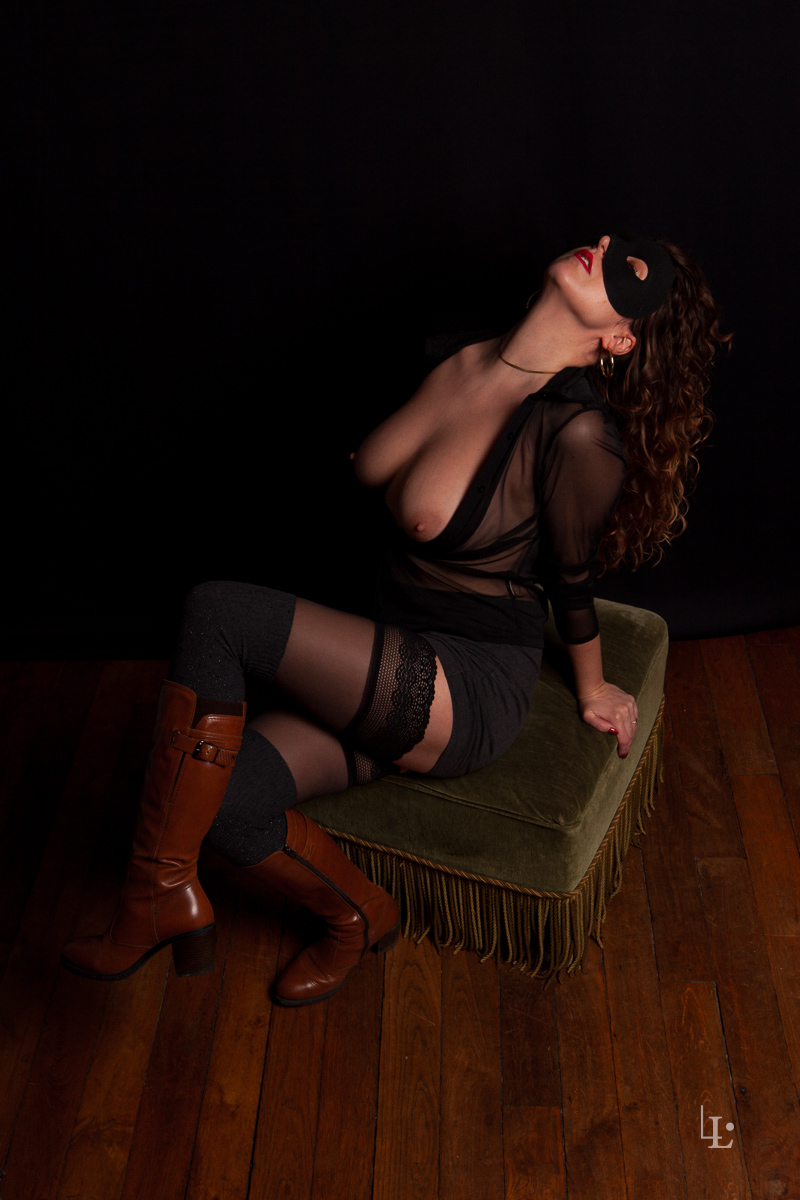 Une jeune femme pose en studio poitrine apparente assise sur un pouf. Elle porte des bottes marron et un masque noir.